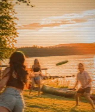 Friends play frisbee on the grass by a lake at sunset, warm and golden on SUNDAY.